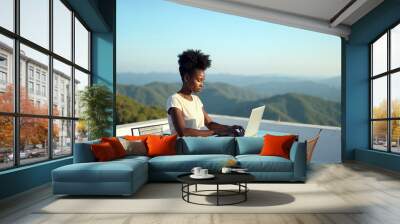 A Black female sitting at a glass table on a rooftop, typing on a laptop, with a panoramic view of rolling hills and a clear blue sky Wall mural