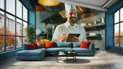 A cheerful famous chef uses a tablet for recipes while working in a modern kitchen. Wall mural