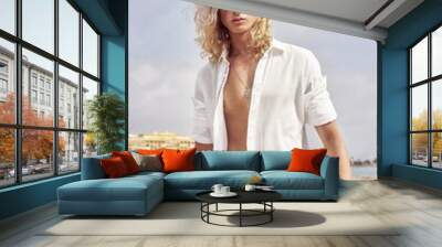 Vertical shot of a young handsome male with long blond hair wearing an unbuttoned shirt at the beach Wall mural