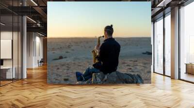 musician sitting on the rock and playing saxophone at sunset in the desert Wall mural