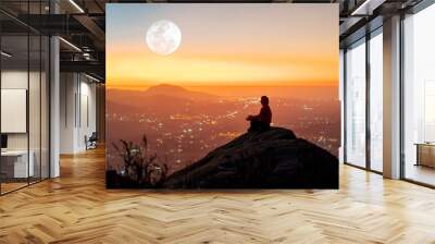 person silhouette sitting on the top of the mountain meditating over the city light in the valley at sunset. Wall mural