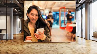 young latin woman holding a cell phone in her hands sitting and smiling in a cafe  Wall mural