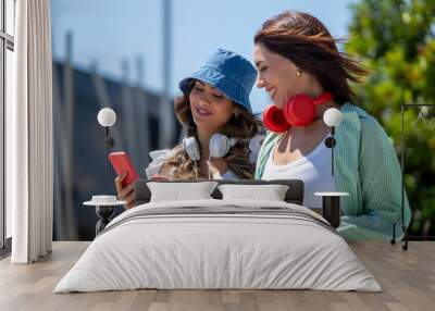 Two Women Smiling and Looking at a Smartphone Together Outdoors, Both Wearing Headphones and Casual Summer Outfits on a Sunny Day. Wall mural