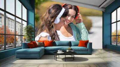 Two Women Smiling and Looking at a Smartphone Together Outdoors, Both Wearing Headphones and Casual Summer Outfits on a Sunny Day. Wall mural