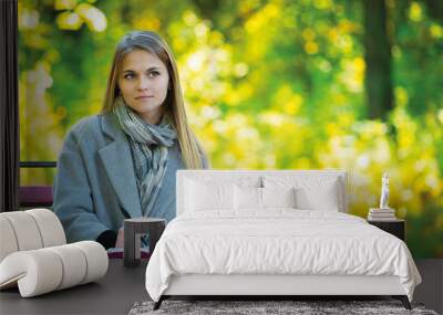 Young blonde girl with umbrella sitting on a park bench in the autumn season. girl in a gray coat drinks coffee, looks into a tablet, talks on the phone, reads a book, writes in a notebook Wall mural