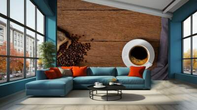 Top view above of Hot fresh black coffee with milk foam in a white ceramic cup with coffee beans roasted in sack bag on wood table background. Flat lay with copy space. Wall mural