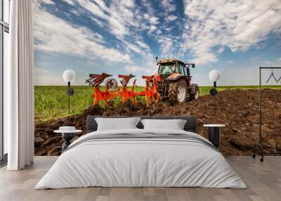 Red tractor is engaged in plowing the soil in a vast green farmland under a cloudy sky Wall mural