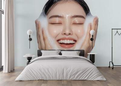 A beautiful korean woman with her eyes closed, smiling and washing her face in the morning with a white background. Her skin is glowing from facial care products. Wall mural