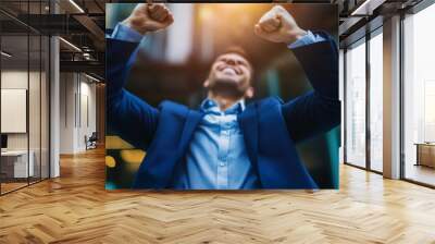 A joyful businessman celebrates success with raised arms, embodying triumph and positivity against an urban backdrop. Wall mural