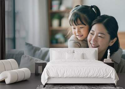 Asian woman sitting on the sofa with her daughter into hugging mom's shoulder, both smiling happy on living room. Wall mural