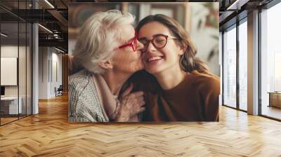 Concept of a happy senior woman with glasses and her adult daughter hugging and kissing in the living room of their home, celebrating Mother's Day. Wall mural