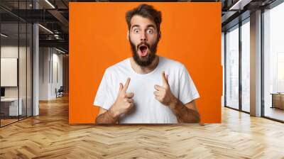 Portrait of astonished young bearded man wearing white t-shirt standing with open mouth pointing away at advertisement area, copy space, close-up, isolated on orange background Wall mural