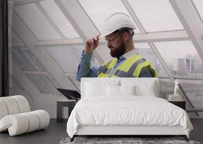 Close up of male chief engineer in hardhat and vest standing inside commercial building construction site using tablet to look at architectural plan of panoramic glass roof. Wall mural