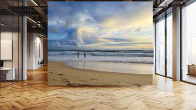 Beach and sunset. Amazing beautiful sky with sun glow and pattern of clouds. Two men stand in the sea in shallow and talk with each other. Clean yellow sand with footprints. Awesome beauty of nature. Wall mural