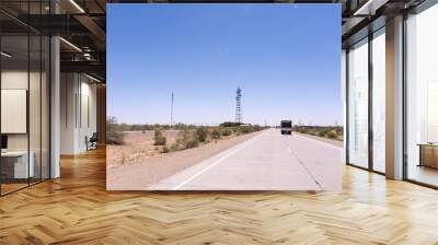 A truck is driving down a road with a tall tower in the background. The sky is clear and blue, and the road is empty Wall mural