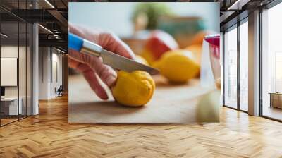 A woman cutting a lemon with a blue knife on a wooden table surrounded by other fruits. Wall mural