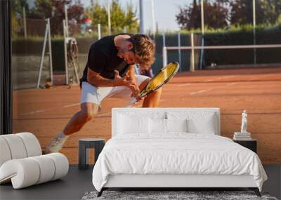 Excited sportive man with racket celebrating triumph on tennis court in bright sunlight looking expressive.   Wall mural