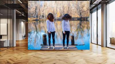 Young twin sisters in same clothes shaking hair at lake on autumn day in the forest Wall mural