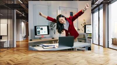 Asian businesswoman celebrates success in her office at night, raising her arms in front of her laptop, feeling happy and excited about her work Wall mural