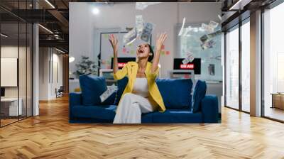 Businesswoman celebrates success by throwing money in the air on a blue sofa in an office, radiating happiness and excitement Wall mural