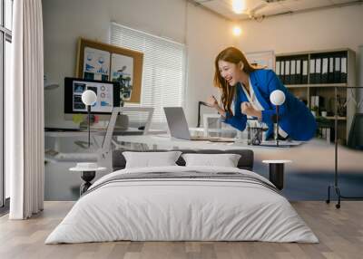 Businesswoman is looking at her laptop and raising her fists in the air, excited about achieving a goal at work. The office is bright and modern Wall mural