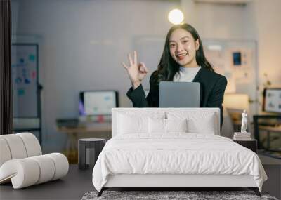 Confident asian businesswoman smiling in modern office, holding laptop and making ok sign gesture, exuding success and happiness in work environment Wall mural