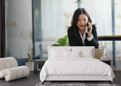 Happy smiling Asian business woman working on laptop at office, using smart phone. Businesswoman sitting at her working place Wall mural