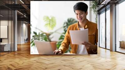 Smiling Asian man working on laptop in modern office space. Wall mural