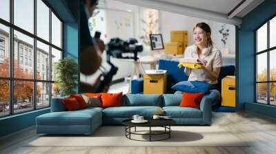 Smiling young asian influencer sitting on sofa and holding clothes while recording video blog about new fashion trends for her followers Wall mural