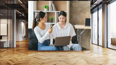 Two young female friends surfing the internet together on a laptop, video conference, work from home concept. Wall mural