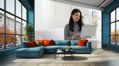 Young beautiful Asian woman using laptop computer while sitting on the sofa at her living room. Wall mural