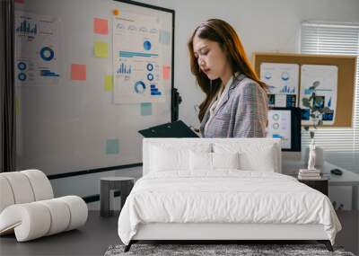 Young businesswoman is standing in an office and reviewing financial data on a clipboard, with charts and graphs displayed on a whiteboard in the background Wall mural