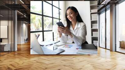 Young happy smiling professional Asian business woman manager, female worker holding cellphone using mobile phone standing in office hall working on smartphone texting message. Wall mural