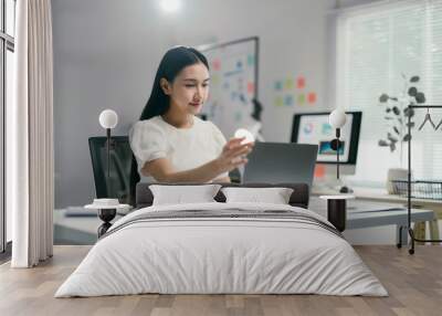 Young professional woman is sitting at her desk and turning on a laptop, ready to start a productive day of work in a modern office Wall mural