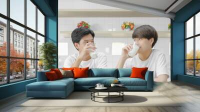 Young asian LGBT gay couple happy drinking milk and eye contact together during breakfast in dining room. Selective focus and copy space. Wall mural