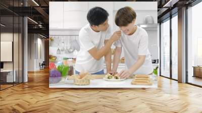 Young asian LGBT gay couple having sweet romantic moment during cooking bread roll salad with happy smiling face in white kitchen at home. Selective focus Wall mural