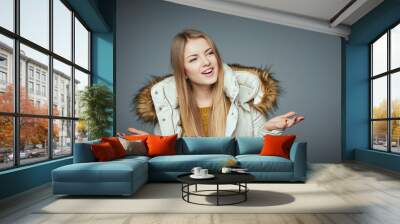 What. Portrait of amazed beautiful girl in winter coat with spreaded hands looking to side on grey studio background Wall mural