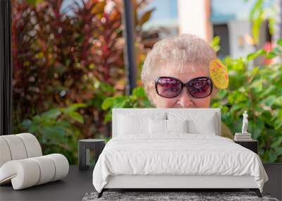 A senior woman at a restaurant in Hawaii, with a cute tropical drink umbrella in her hair. She wears sunglasses. Wall mural