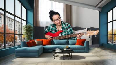 Latin woman sitting on the sofa learning how to play bass with her laptop Wall mural