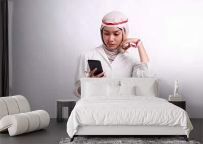 A smart young asian muslim woman with red white flag headband thinking an idea while holding her phone, isolated on white background. Indonesia's independence day. Wall mural