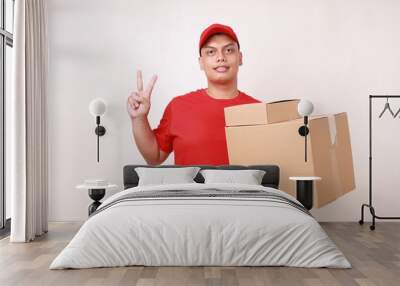 Happy Asian courier man in red holding a cardboard box while showing two fingers. Isolated on white Wall mural