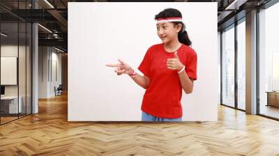 Happy young Asian girl in red white flag ribbon pointing fingers at copy space, showing thumb up gesture isolated on white background. Indonesian independence day on 17 august concept Wall mural