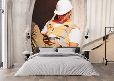 A man in a hard hat is sitting in a hole with his cell phone wearing an orange vest. A construction worker in helmet and vest sits with a coffee and a phone on his lunch hour near a construction site Wall mural