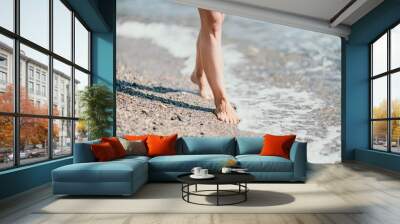 A woman walks along the beach, legs close-up. Barefoot woman standing in sea, summer vacation on beach resort. Naked female legs in transparent calm water Wall mural