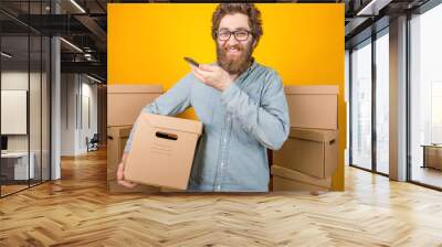 Male delivery man holding a cardboard box in his hands while standing among many boxes with parcels on a yellow background takes an order by phone Wall mural