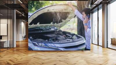 A broken down car, engine open and smoking, serious driver looking at the engine and using smartphone call for service. Wall mural