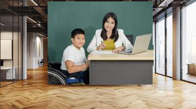 A handicapped boy in a wheelchair listening attentively to the young lady teacher explaining mathematics. Disabled kids classroom, happy disability kid concept.. Wall mural