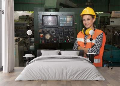 Portrait of confident young female mechanical engineer in a protective uniform, helmet, and sound protector controls the CNC Machinery while working at the plant. Wall mural