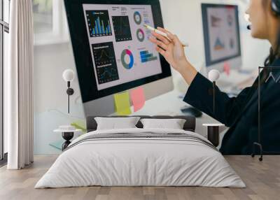 Businesswoman wearing headset is analyzing financial data on computer screen, pointing at charts and typing on keyboard Wall mural