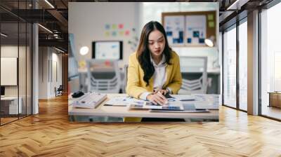 Focused asian businesswoman analyzing data in modern office, surrounded by technology and paperwork, reflecting determination for career success and business growth Wall mural